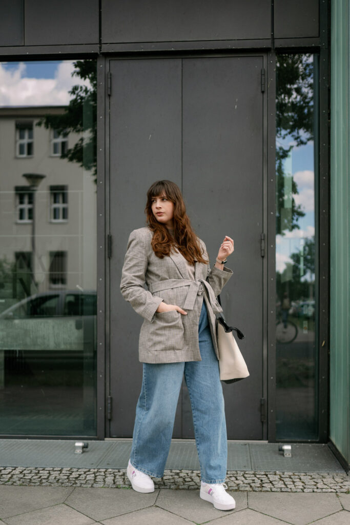Andrea trägt eine weite Jeans mit grauem Oversize-Blazer, weißen Sneakern und cremefarbener Tote Bag – perfekter Casual-Business-Style für den Alltag.