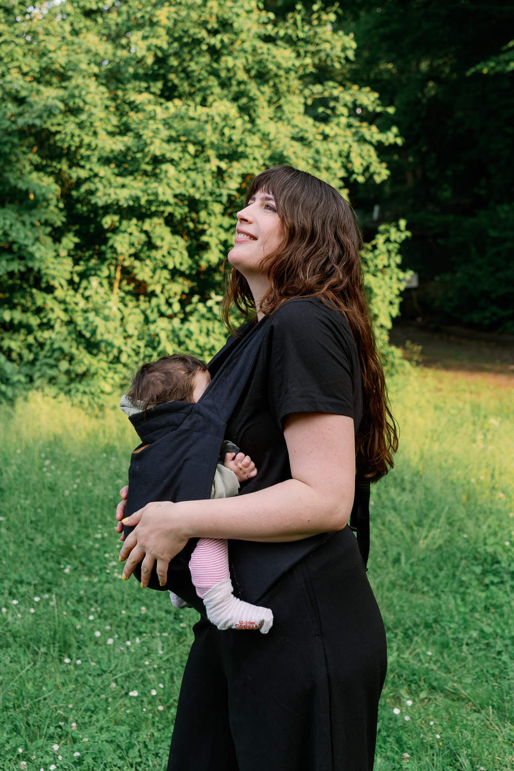 Mama blickt lächelnd nach oben, Baby in Trage – Alltagsroutinen für mehr Wohlbefinden