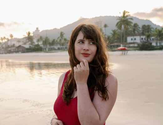 Andrea von andysparkles im roten Badeanzug am Strand bei Sonnenuntergang, lächelnd und mit der Hand am Kinn – bequemer Badeanzug nach der Schwangerschaft