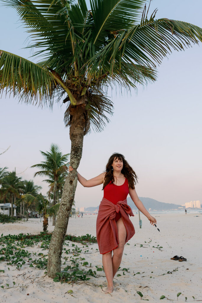 Andrea von andysparkles im roten Badeanzug mit Tuch um die Hüfte lehnt an einer Palme am Strand – Sommerlook nach der Schwangerschaft