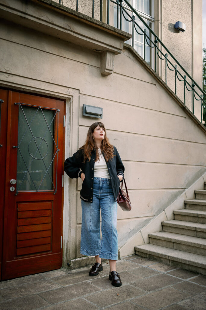 Andrea von andysparkles trägt eine schwarze Collegejacke, weißes Top und weite Jeans im Herbstlook 2025, kombiniert mit einer bordeauxfarbenen Tasche.