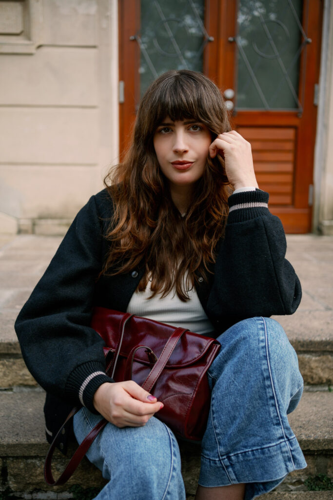 Herbsttrend 2025: Andrea sitzt in schwarzer Collegejacke und Denim-Jeans auf den Stufen, dazu eine glänzende bordeauxrote Handtasche als Farbakzent.