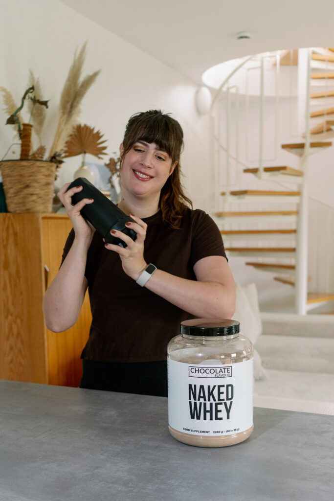 Andrea gießt sich einen Schoko-Proteinshake aus dem Shaker ins Glas, das Grass-Fed Whey Protein von Naked Nutrition steht auf dem Tisch.