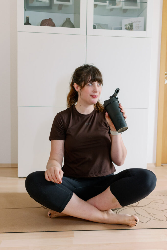 Andrea sitzt auf der Yogamatte, trinkt ihren Proteinshake und lächelt – ruhiger Moment nach einer kleinen Workout-Einheit.