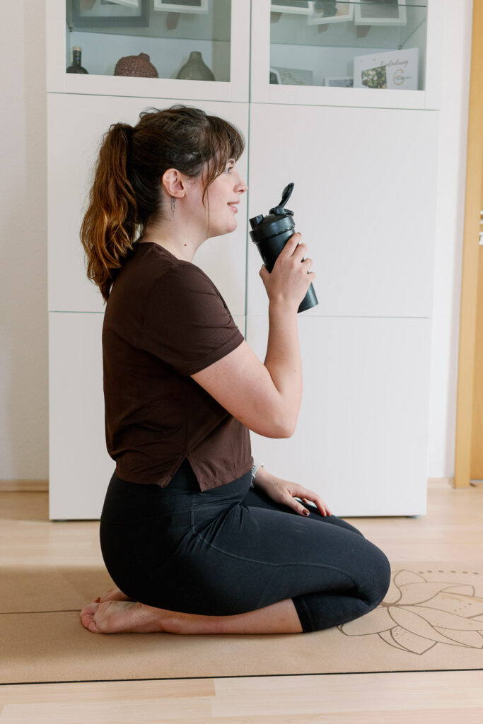 Andrea sitzt auf der Yogamatte, trinkt ihren Proteinshake und lächelt – ruhiger Moment nach einer kleinen Workout-Einheit.