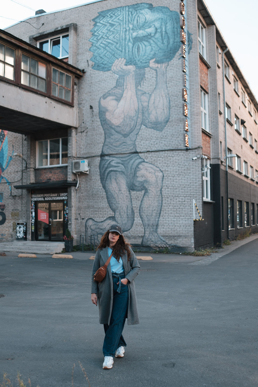 Streetstyle mit Jeans im Winter: Andrea spaziert durch das Telliskivi-Viertel in Tallinn, trägt eine Paperbag-Jeans, grauen Mantel und Sneaker vor urbaner Streetart-Kulisse.