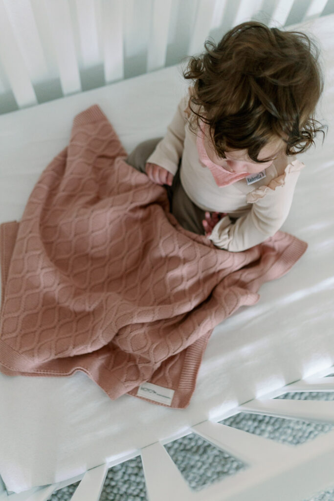 Baby sitzt mit rosa Strickdecke im weißen Babybett vor der Abendroutine