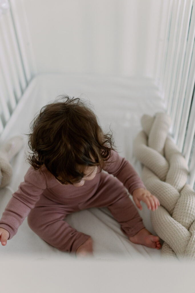 Baby sitzt im Gitterbett im rosafarbenen Outfit und spielt am Nestchen
