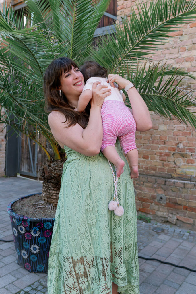 Andrea von andysparkles hält ihr Baby im Arm im Garten vor Palmen und Backsteinwand, grünes Spitzenkleid