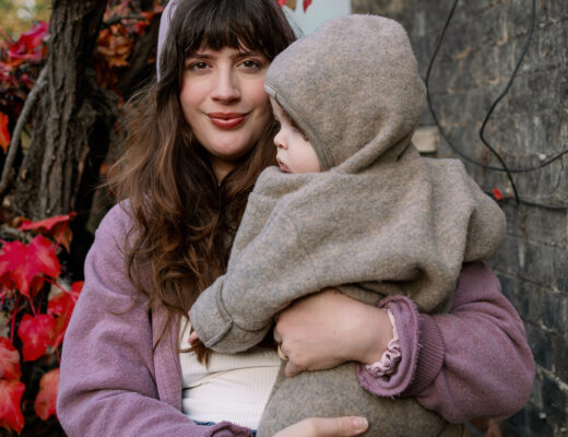 Stil als Mama im Herbst – Porträt einer Mama mit Baby im Wollanzug vor roten Herbstblättern