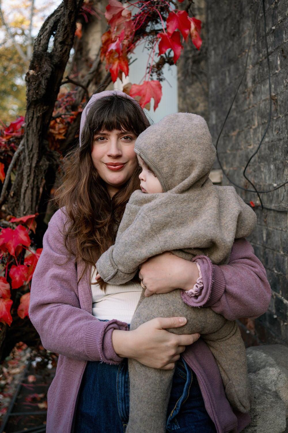 Stil als Mama im Herbst – Porträt einer Mama mit Baby im Wollanzug vor roten Herbstblättern