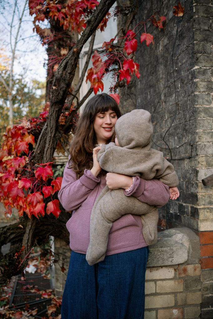 Mama hält ihr Baby im Wolloverall im herbstlichen Setting an Backsteinwand