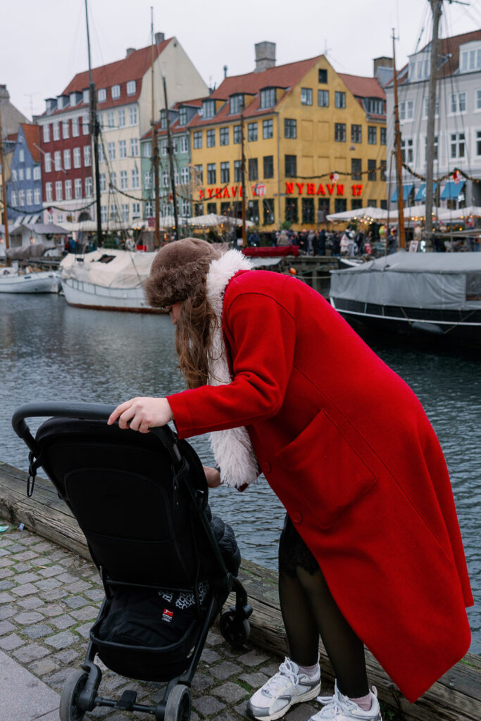 Andrea von andysparkles im roten Mantel beugt sich über den Kinderwagen an Nyhavn in Kopenhagen, Virale Winter Fashion Hacks mit Baby und Fußsack bei Wind