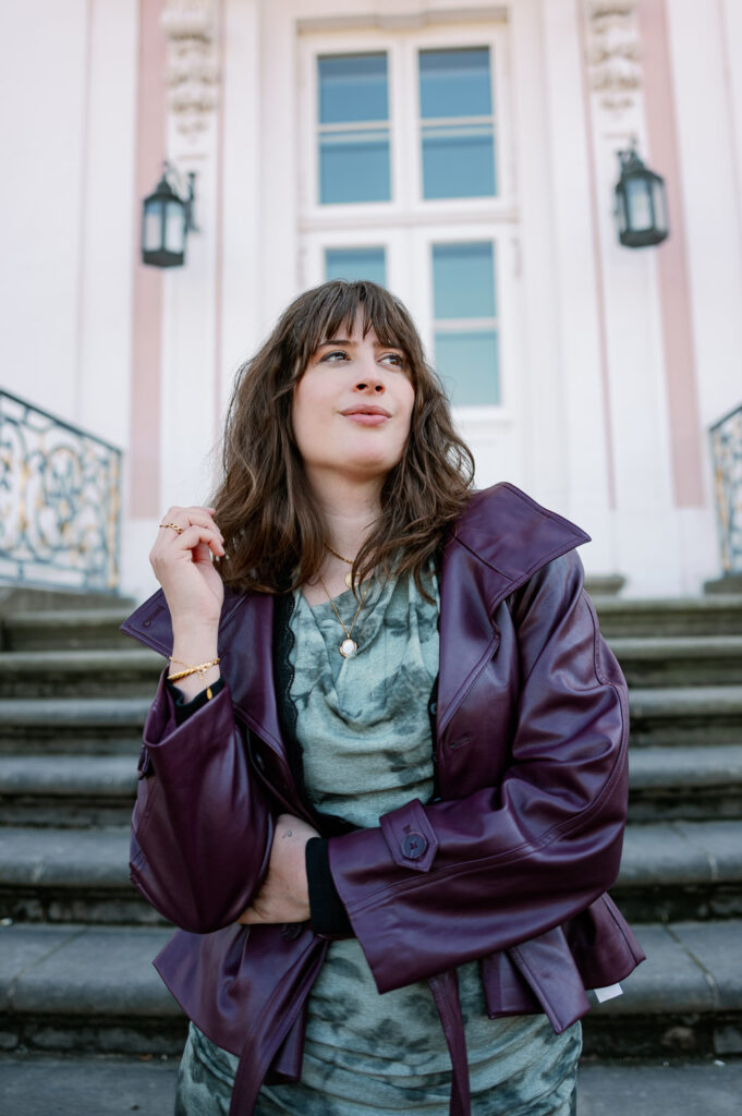 Andrea von andysparkles in bordeauxfarbener Lederjacke und grünem Kleid vor einer historischen Fassade