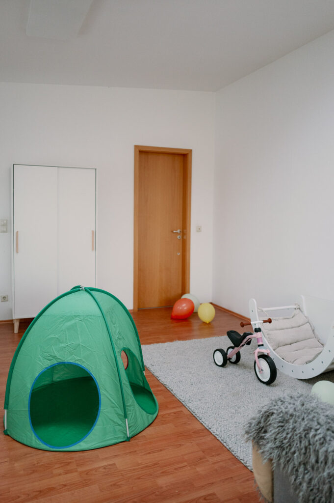 Spielbereich im Kinderzimmer mit grünem Spielzelt, Kleiderschrank, grauem Teppich, Laufrad und Kletterbogen