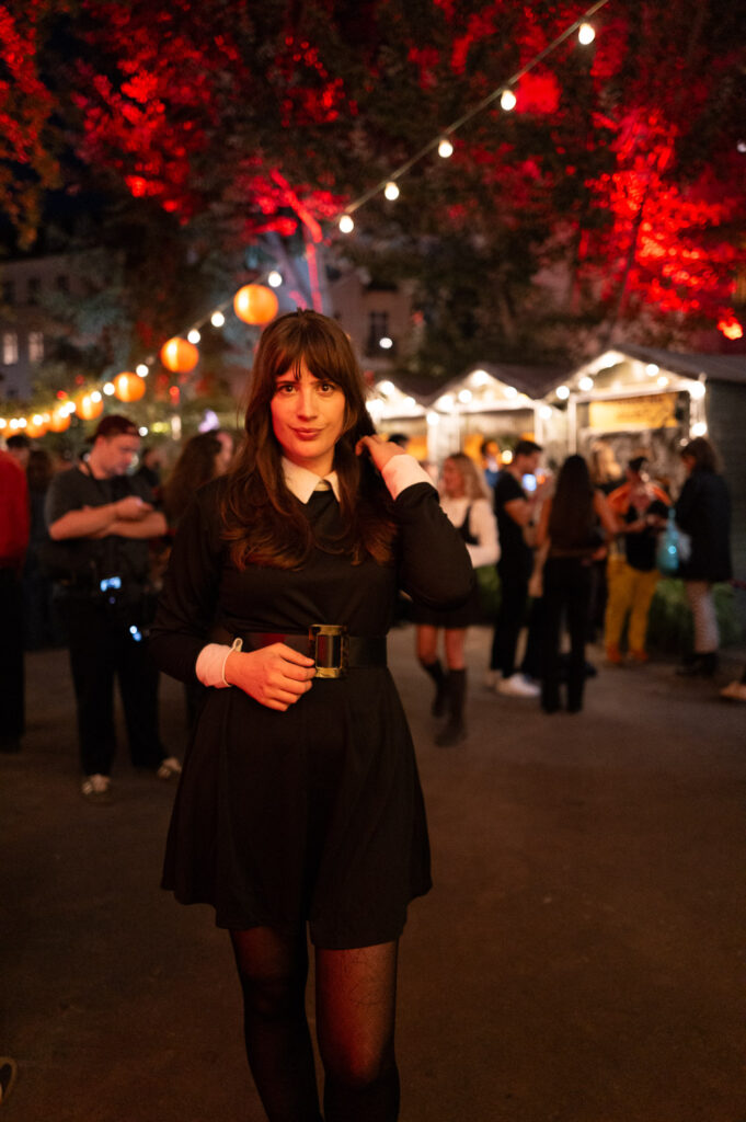 Andy im Wednesday Addams Look mit schwarzem Kleid, weißem Kragen und breitem Gürtel auf einer Halloween-Party unter roten Lichtern
