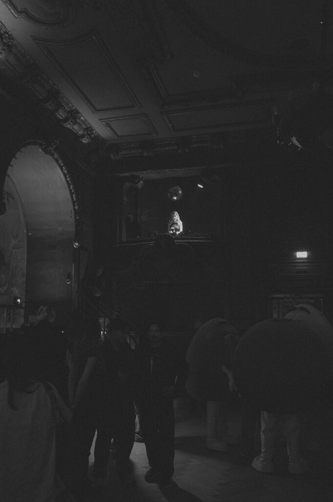 Schwarz-Weiß-Foto einer Halloween-Party in einem dunklen Saal mit Stuckdecke, DJ auf der Empore und Gästen in Kostümen