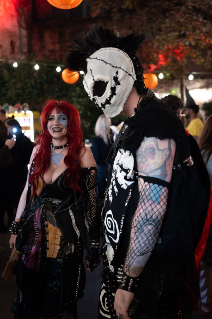 Halloween-Party bei Nacht: Frau mit roter Perücke und genähtem Patchwork-Kleid neben einer Person mit Totenkopfmaske aus Stoff