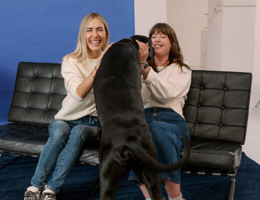 Andrea von andysparkles und Alena Gerber lachen auf einer schwarzen Lederbank, während eine schwarze Dogge zu ihnen hochkommt